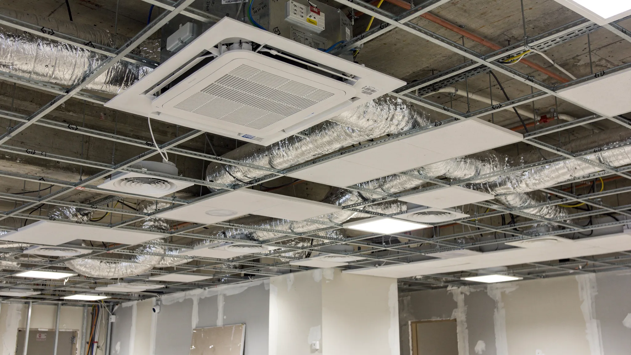 A commercial office false ceiling under installation showing a ductable indoor unit connected to insulated ducts, with visible diffusers and return air grille positions marked along the ceiling grid.