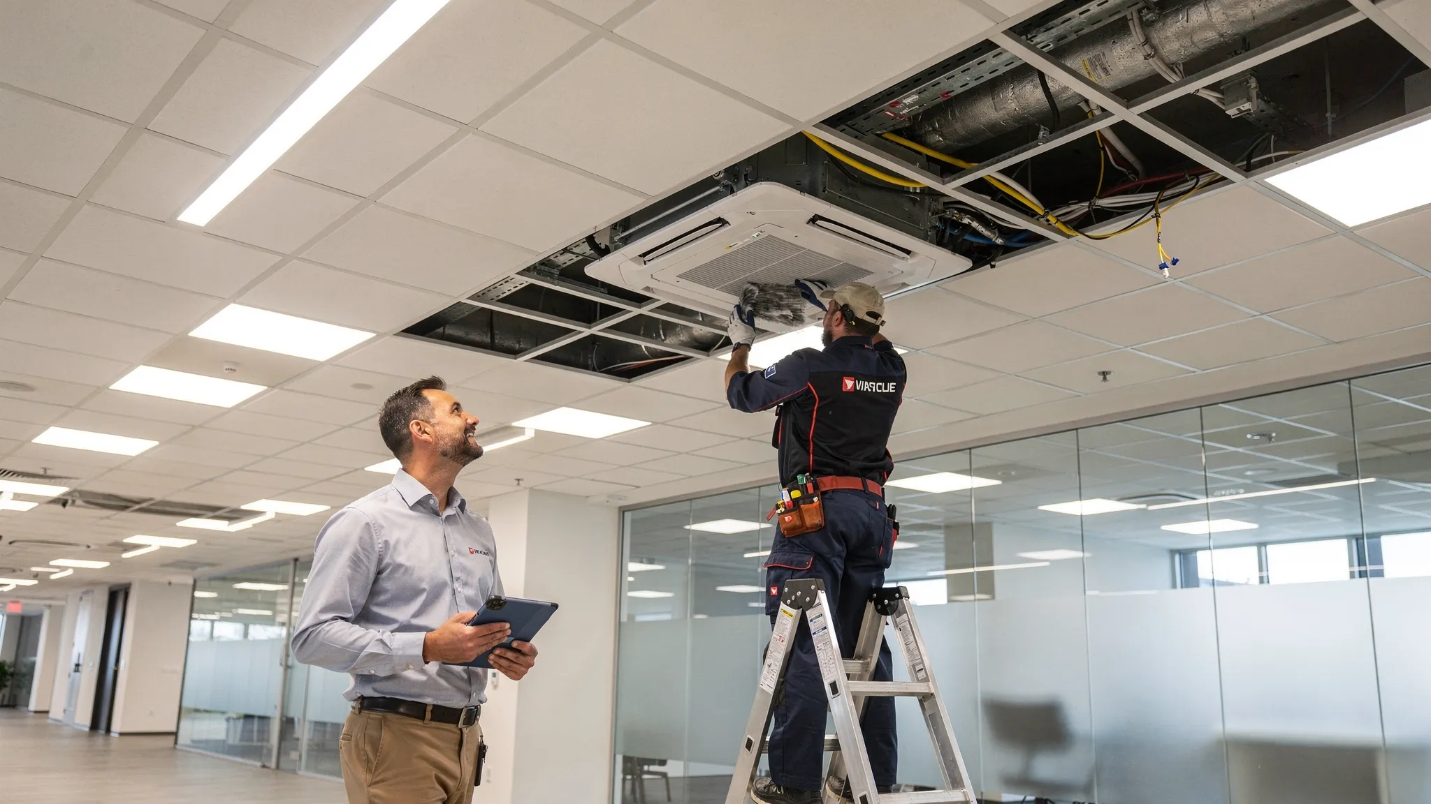 A facility manager walking through a modern office ceiling grid area with an HVAC technician inspecting a VRF indoor unit access panel and cleaning filters, showing planned maintenance and easy service access.