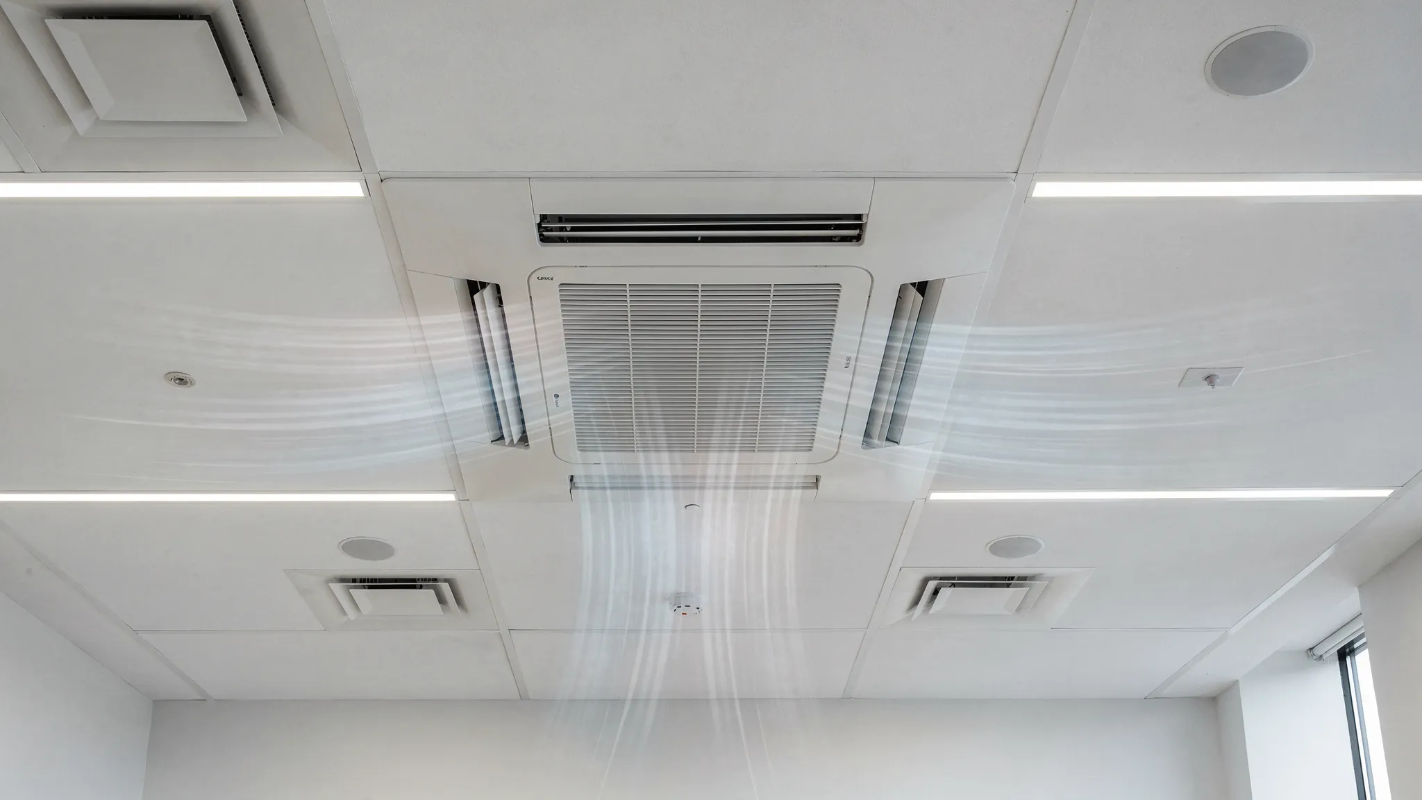An indoor office ceiling showing a cassette AC unit centered in a false ceiling grid with air vents, lighting panels and clear airflow direction lines, representing zoned VRF comfort.